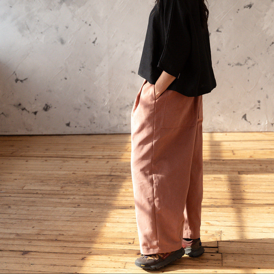 Woman wearing a black top and pink pants standing against a textured wall.