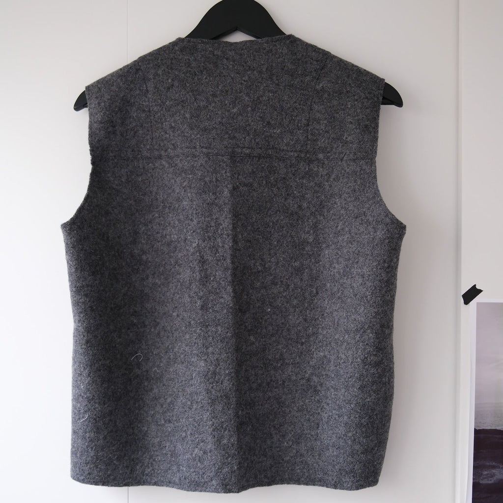 A dark gray textured wool vest hanging on a black hanger against a white wall.