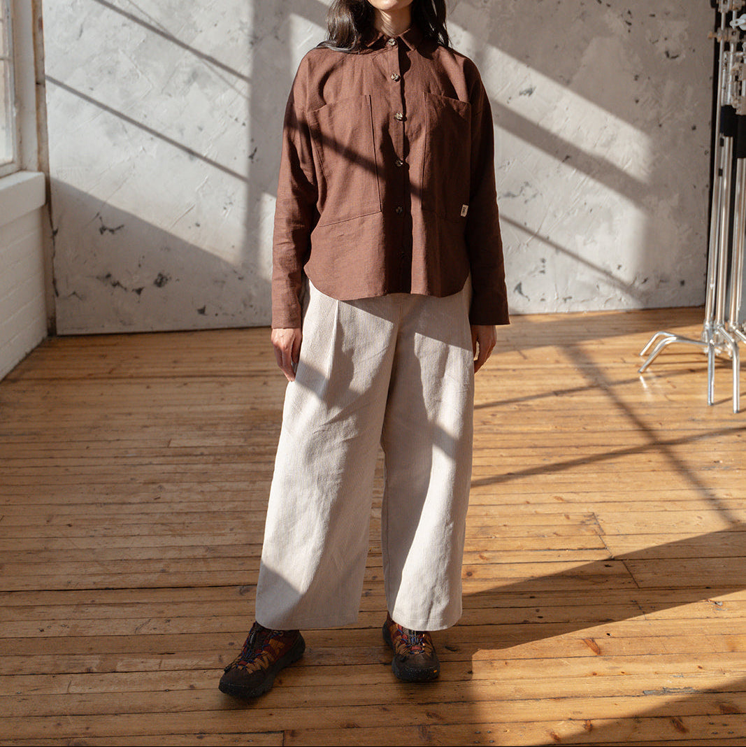 Person wearing a brown shirt and beige pants standing in a sunlit room with wooden floor and textured white wall.