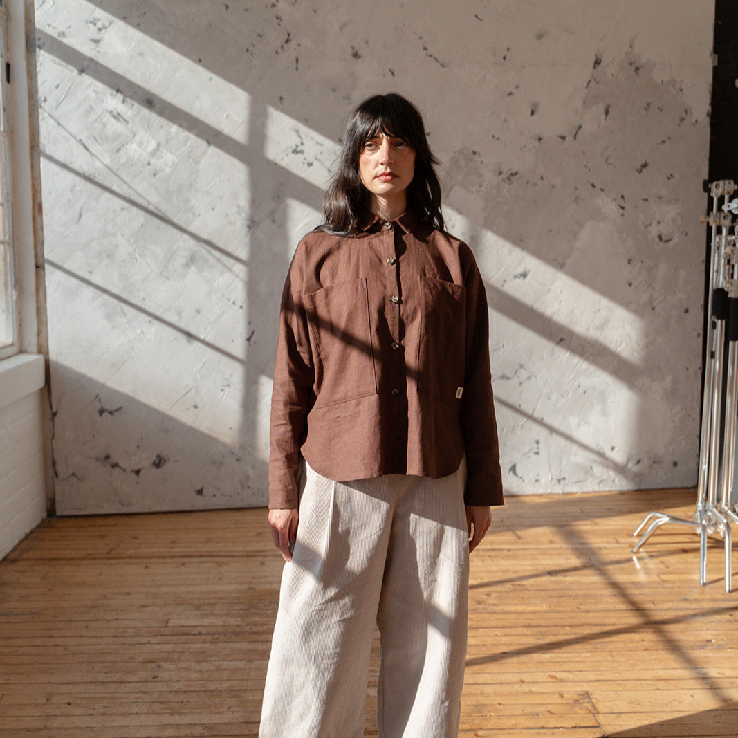 Person wearing a brown shirt and beige pants standing in a sunlit room with wooden floor and textured white wall.