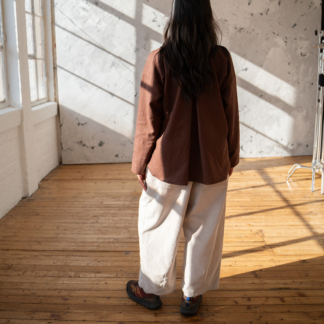 Person standing in a room with wooden floor and white walls, wearing a brown jacket and white pants.