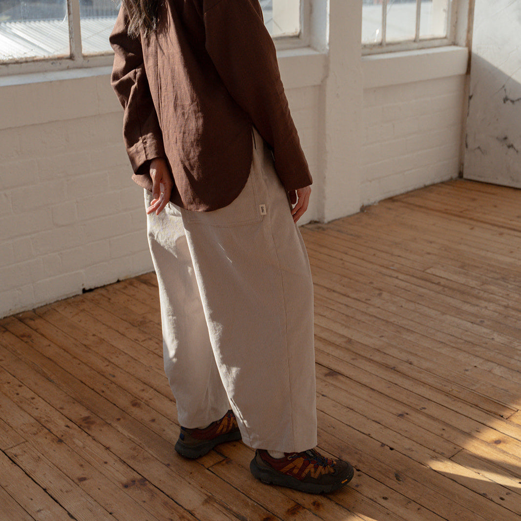 Person standing in a sunlit room with large windows, wearing a brown jacket and white pants.