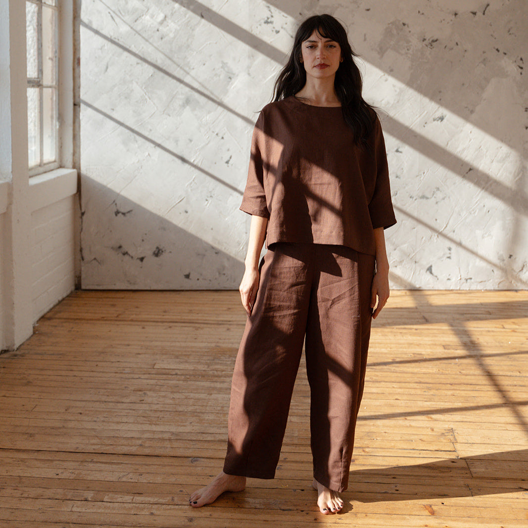 Woman in brown outfit standing in a sunlit room with wooden floor and white walls.