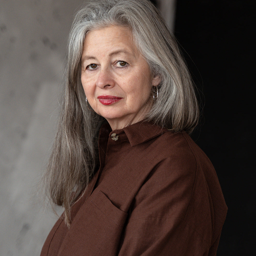 Woman with gray hair wearing a brown shirt against a textured wall.