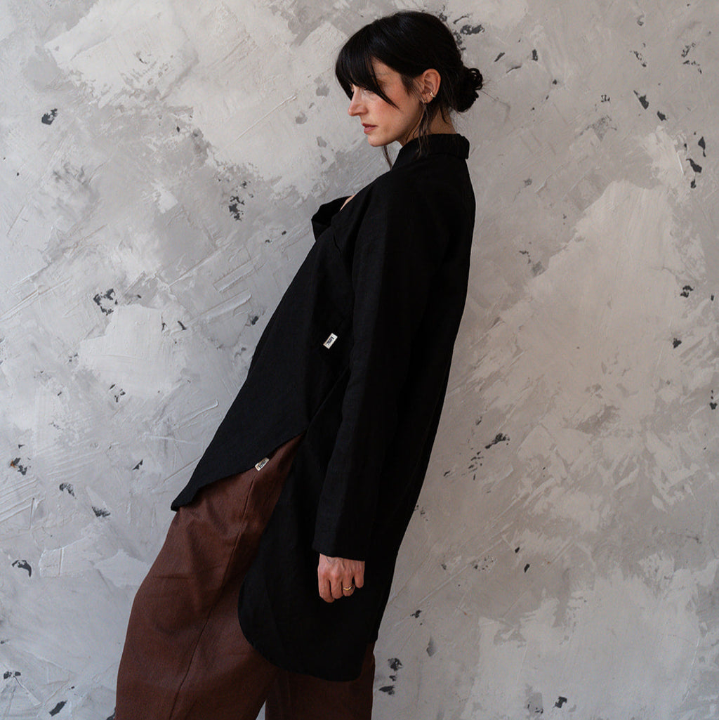 Person wearing a black coat and brown pants against a textured white wall