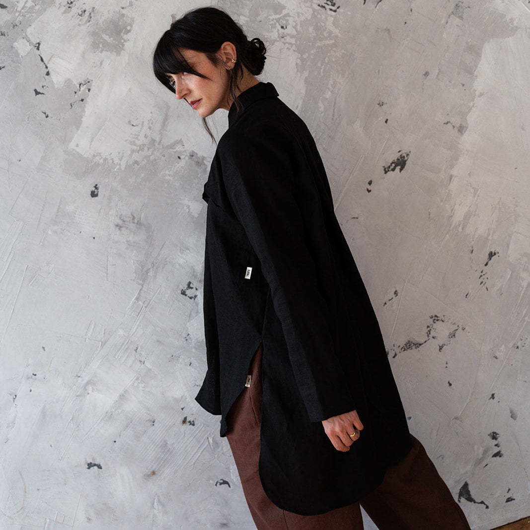 Person wearing a black coat and brown pants against a textured white wall.
