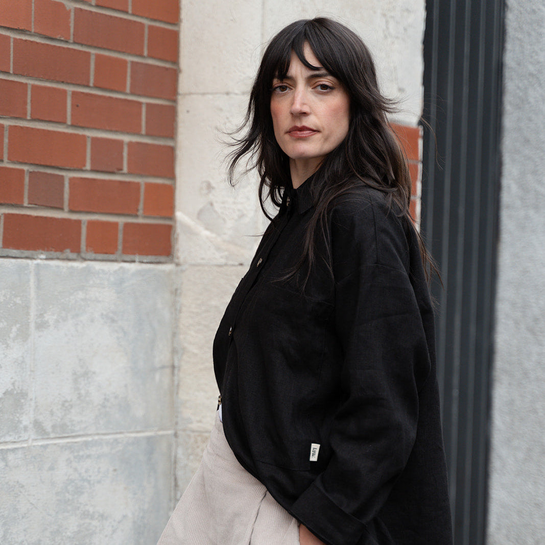Woman wearing a black coat and light-colored pants standing against a brick wall.