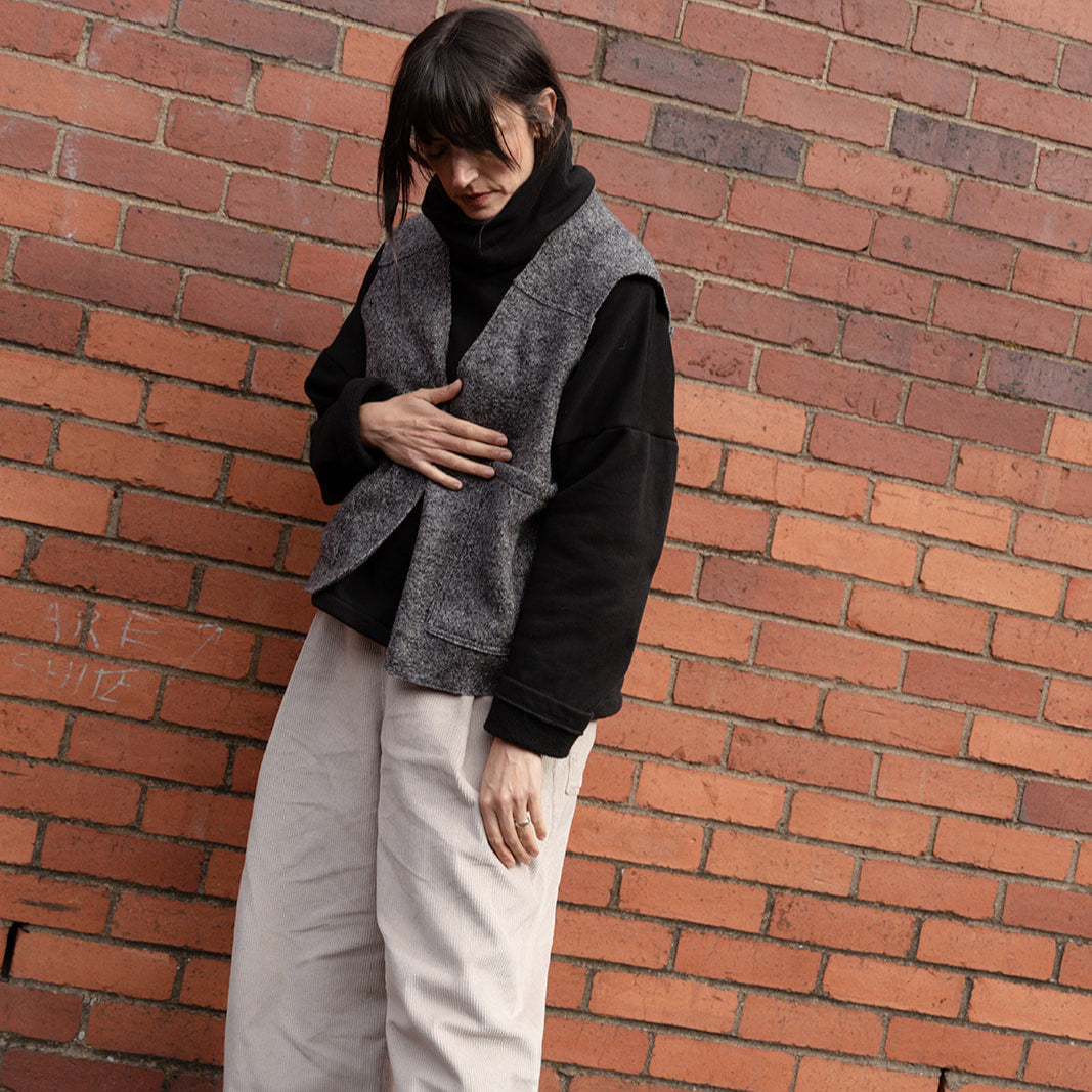 Person wearing a black sweater, gray vest, and white pants against a brick wall.