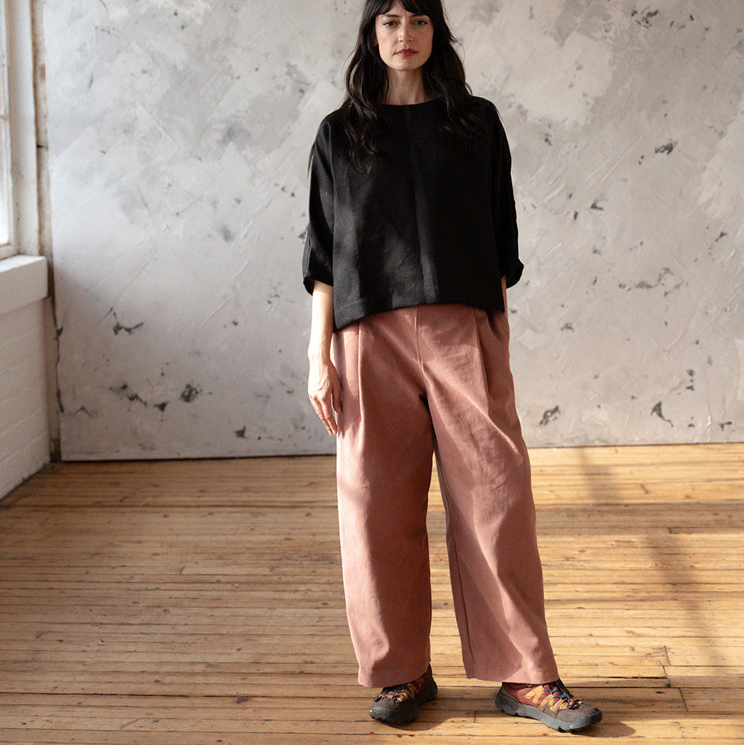 Woman wearing a black top and pink pants standing in a room with wooden floor and white walls.