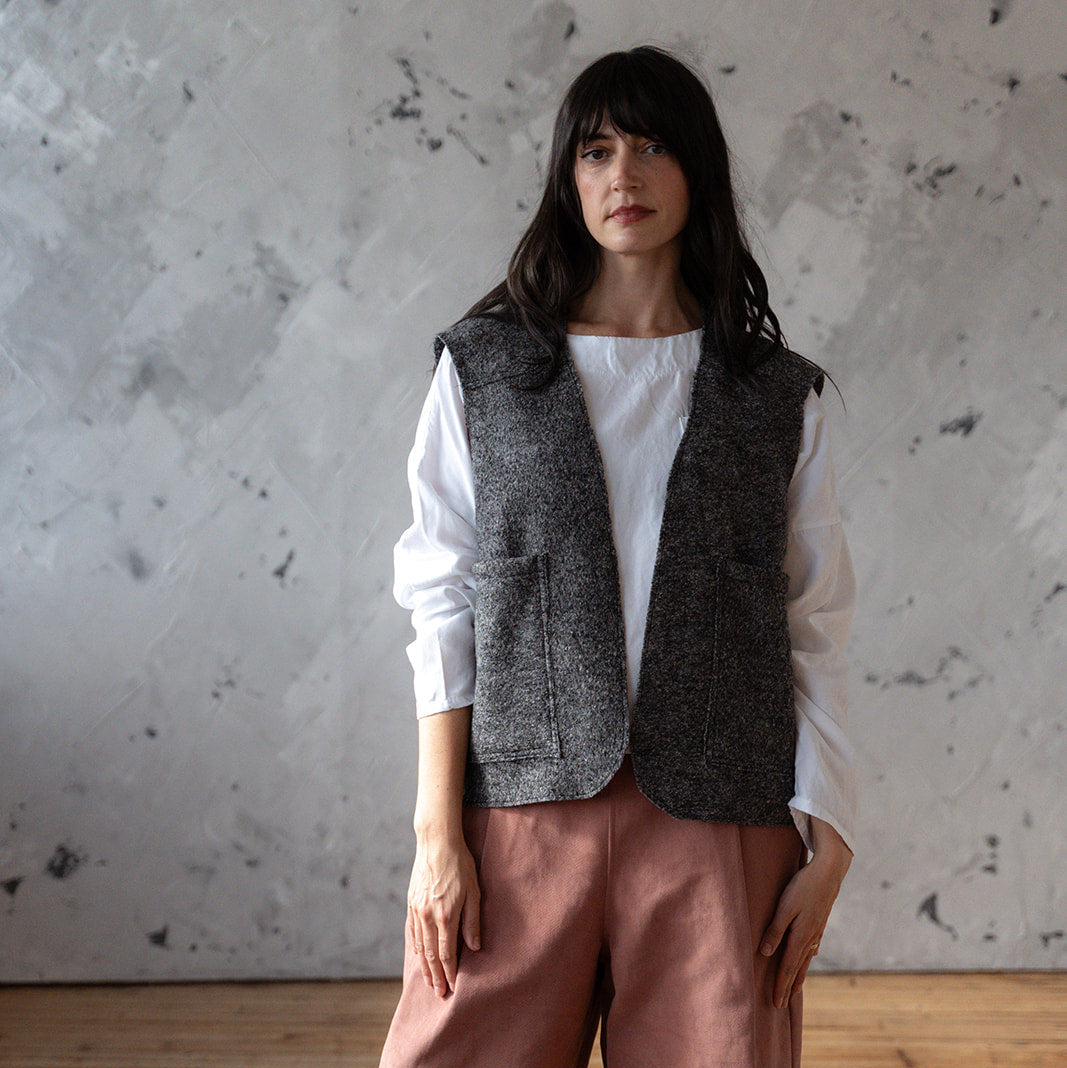 Woman wearing a white shirt, gray vest, and pink pants against a textured wall.