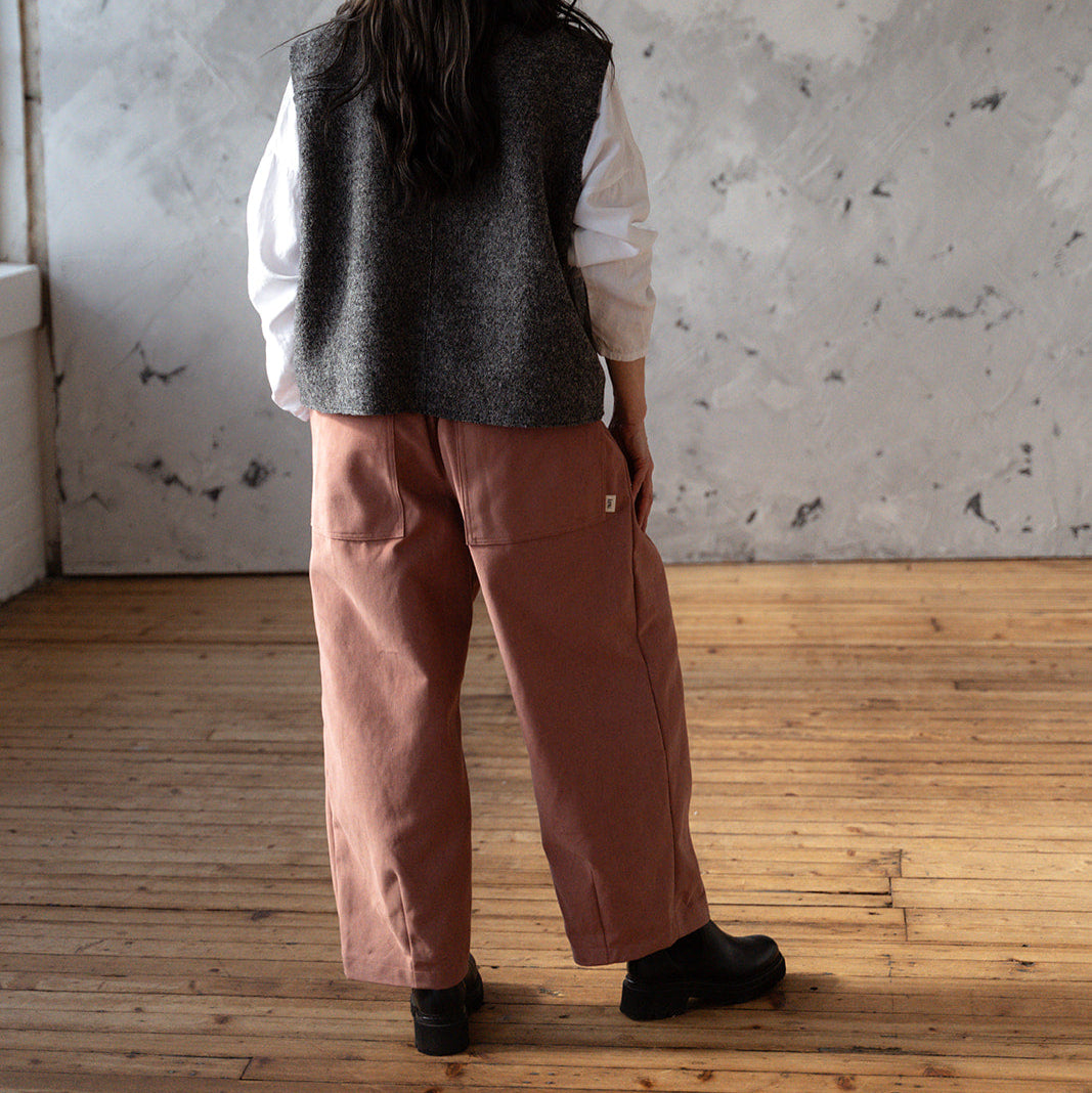 Person wearing a gray vest and brown pants standing in a room with a textured wall and wooden floor.