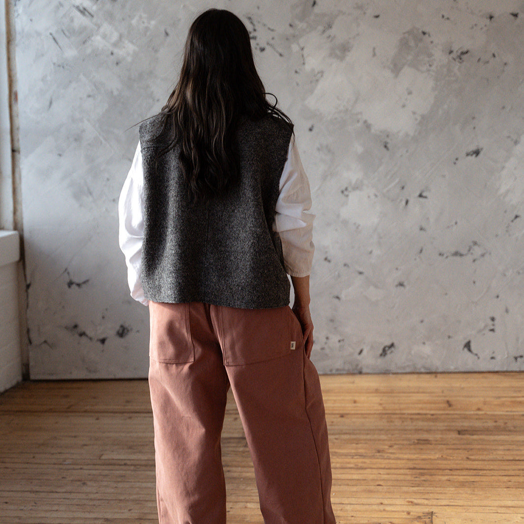 Person wearing a gray vest and brown pants standing in a room with a textured wall and wooden floor.
