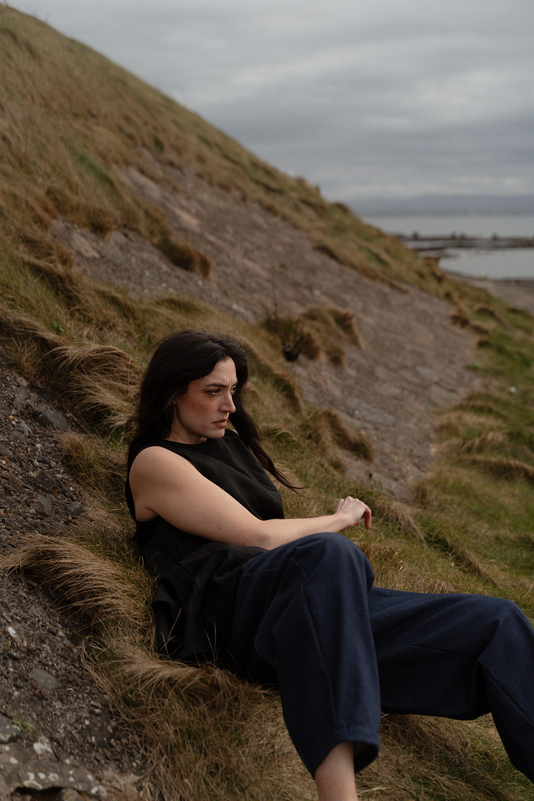 Woman sitting on a grassy hillside with a body of water and cloudy sky in the background