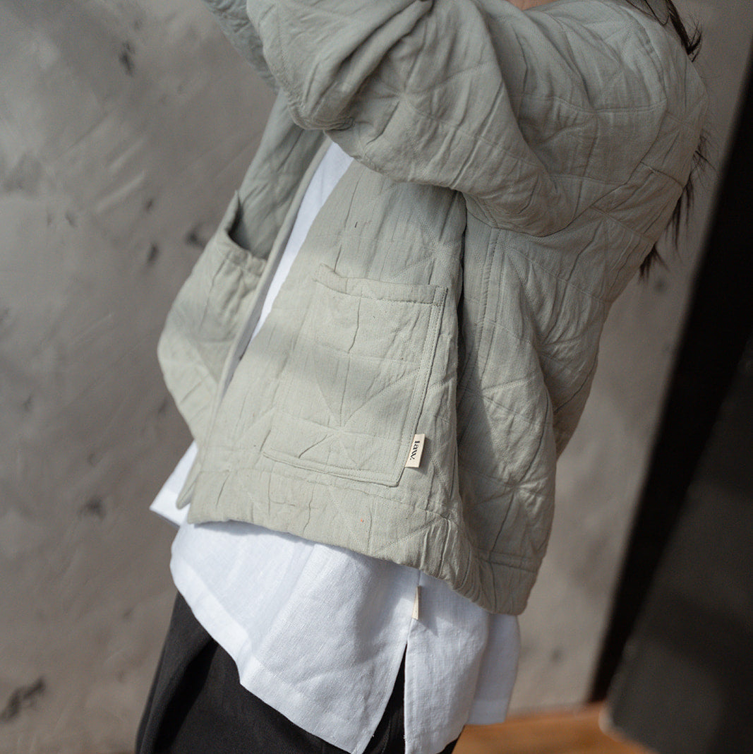 Person wearing a light green textured jacket over a white shirt and dark pants in an indoor setting.
