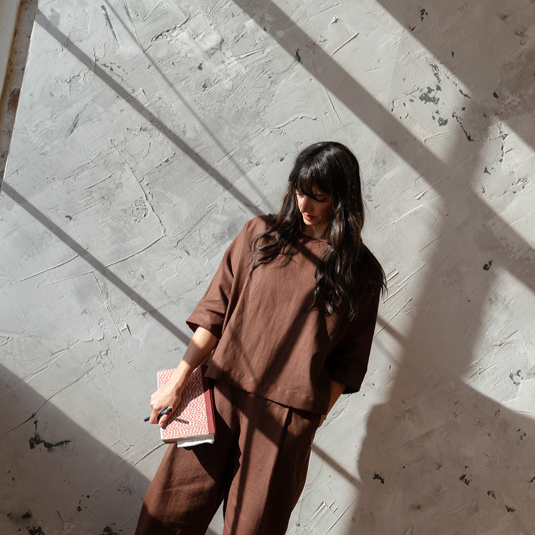 Woman in brown outfit holding a pink book against a textured wall.