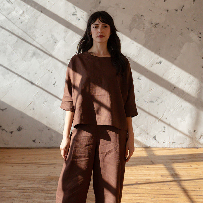 Woman in brown outfit standing in a sunlit room with wooden floor and white walls.