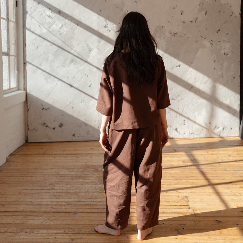 Person wearing a brown outfit standing in a sunlit room with wooden floor and white walls.