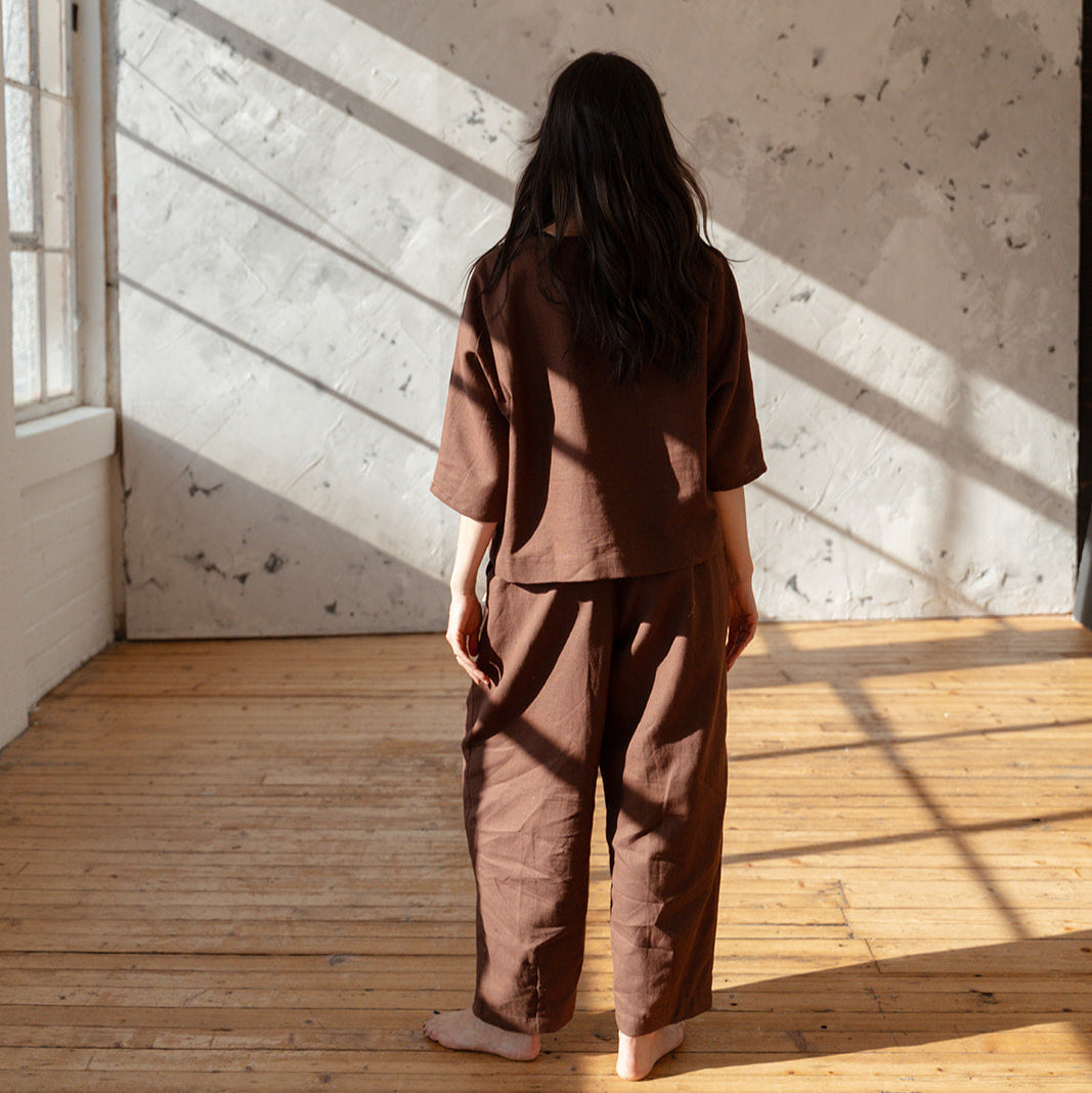 Person wearing a brown outfit standing in a sunlit room with wooden floor and white walls.
