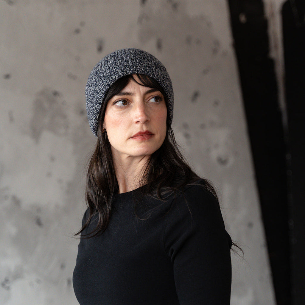 Woman wearing a gray knit beanie and black long-sleeve shirt against a textured wall.