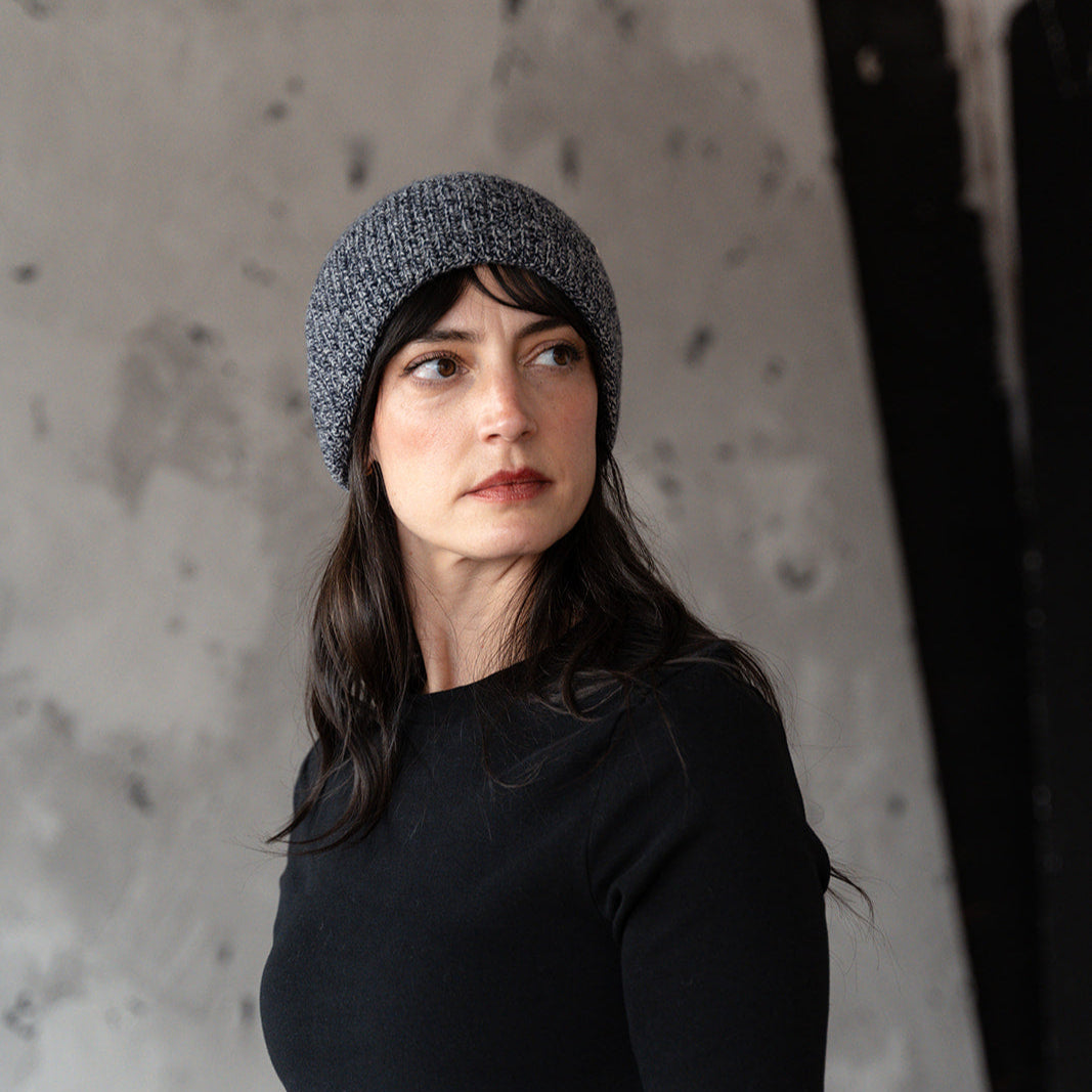Woman wearing a gray knit beanie and black long-sleeve shirt against a textured wall.