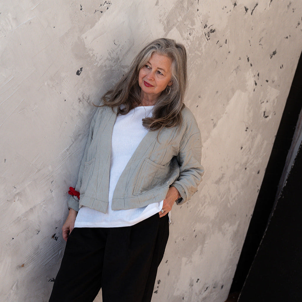 Woman standing against a textured wall wearing a light gray jacket, white shirt, and black pants.