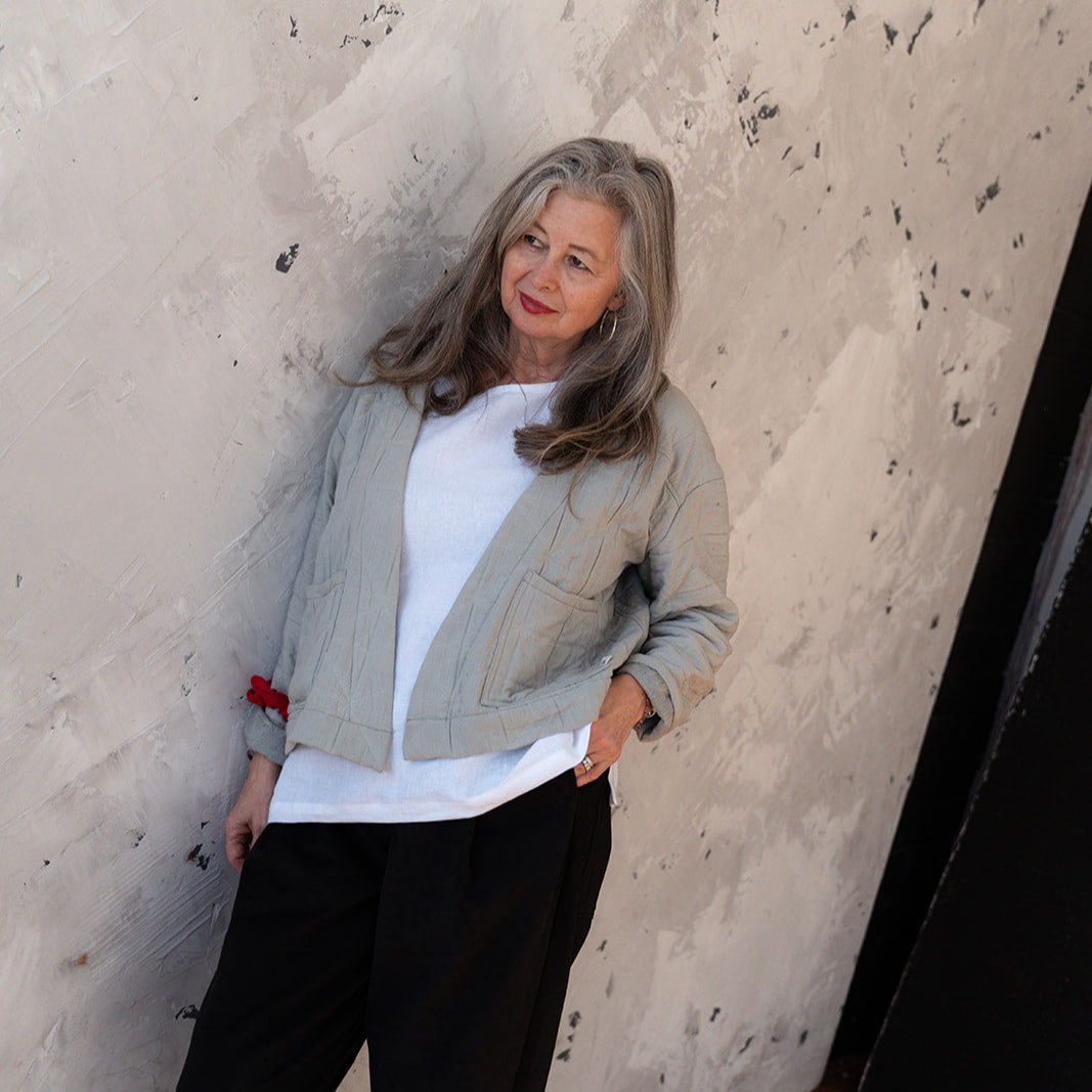 Woman standing against a textured wall wearing a light gray jacket, white shirt, and black pants.