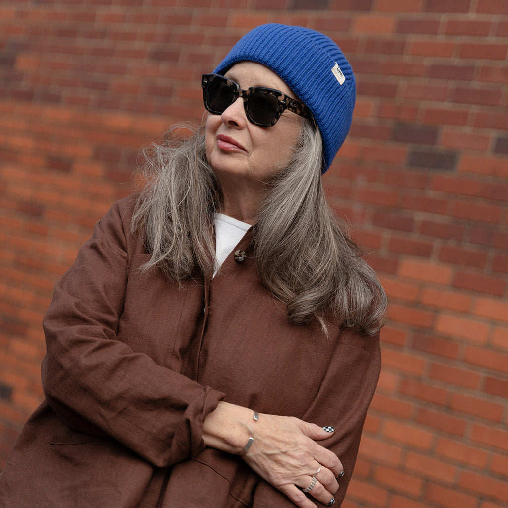 Person wearing a blue beanie and sunglasses, sitting against a brick wall.