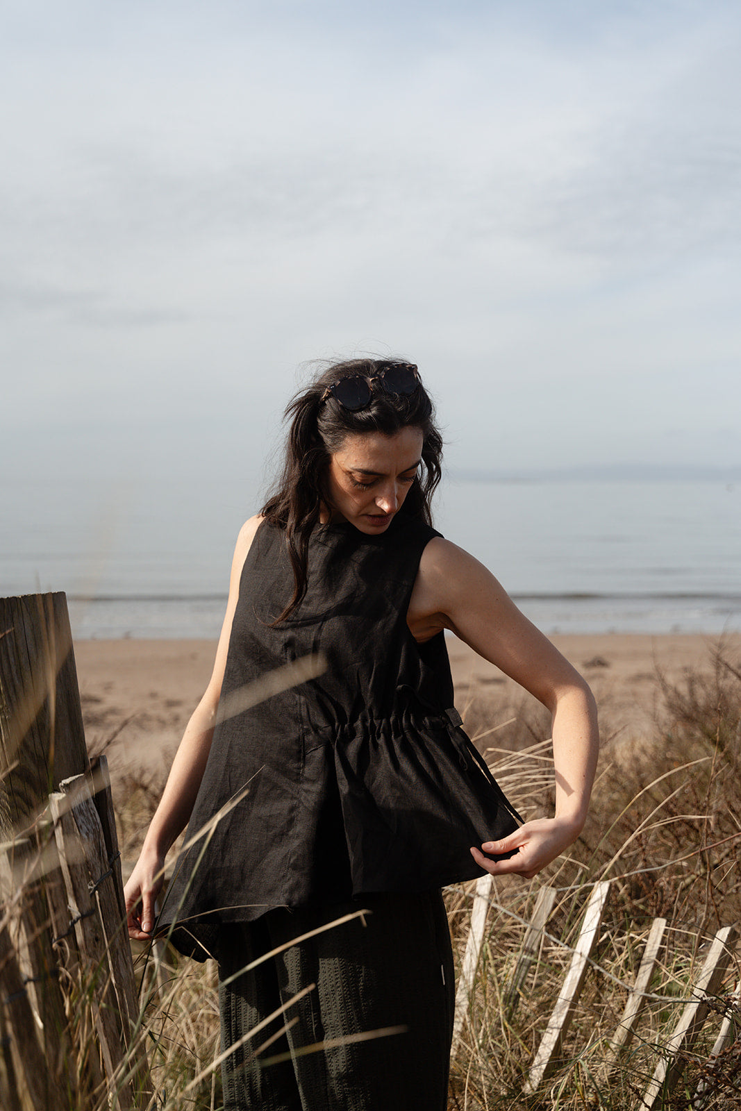 Woman in a black sleeveless dress standing on a beach with ocean in the background