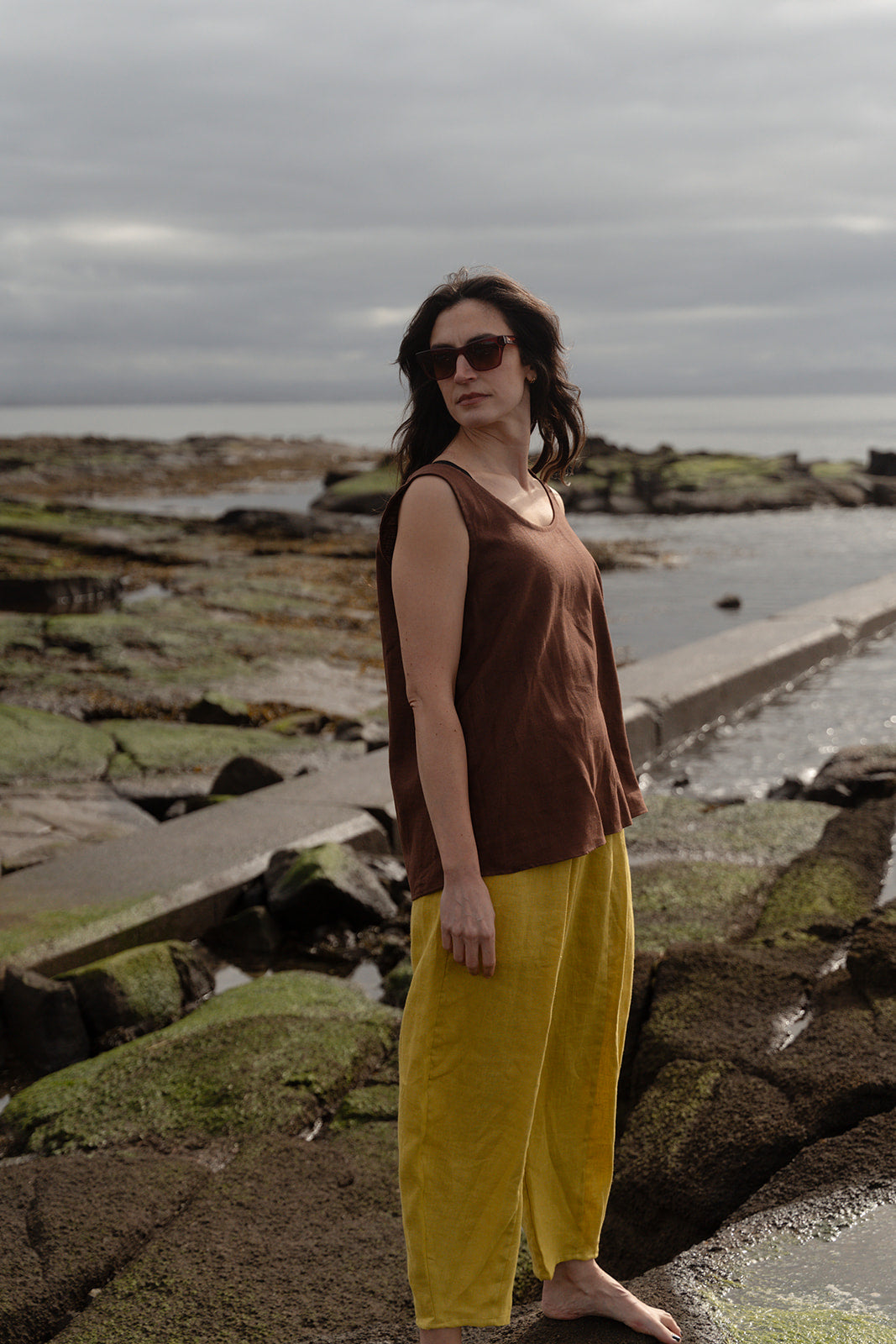 Woman standing on a rocky shoreline wearing a brown top and yellow pants.